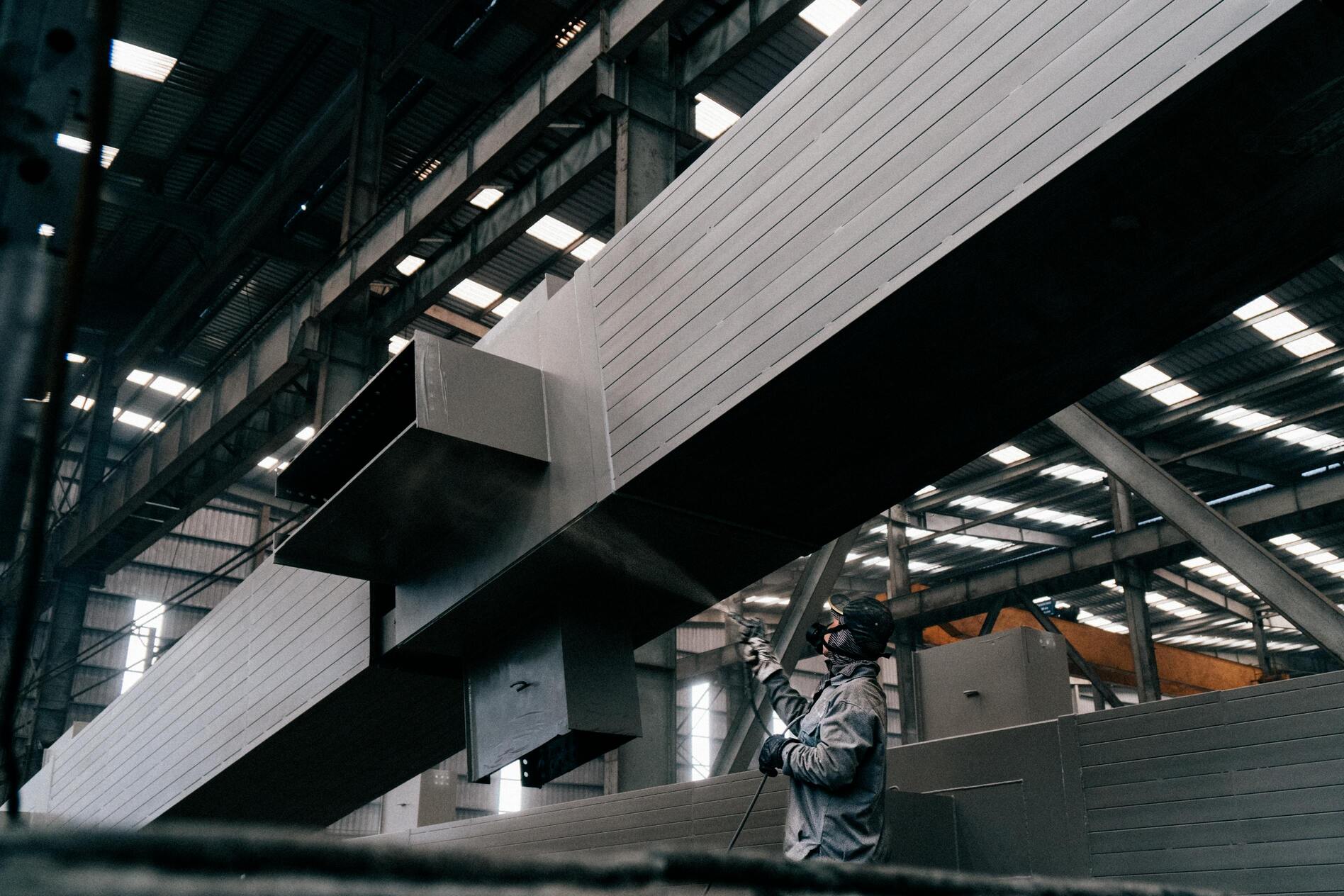 Industrial worker spray-painting steel beams inside a large manufacturing warehouse